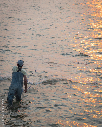 Unidentified Asian fisher man with waterproof wader and hat throwing a cast net at sunrise
