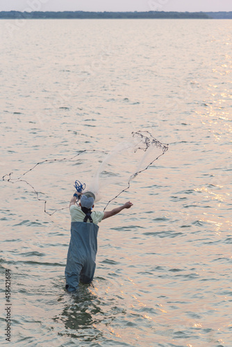 Unidentified Asian fisher man with waterproof wader and hat throwing a cast net at sunrise