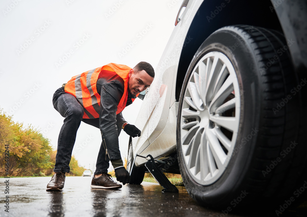 Roadside assistance worker raising vehicle with car jack before changing  flat tire. Young man using special device for lifting car while repairing  wheel on the road. Concept of emergency road service. Stock