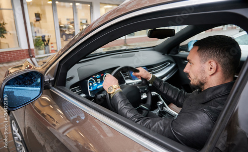 Close-up view of confident, bearded man testing new car in the courtyard of car dealership. Guy putting his hands on the steering wheel and looking straight ahead. © anatoliy_gleb