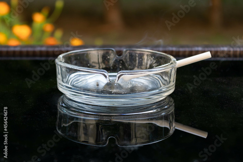 Close up of a transparent ashtray with a lit cigarette reflected on a black glass table