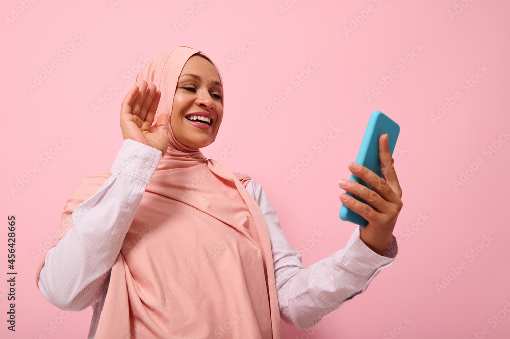 Muslim cute woman of Middle Eastern or Arab ethnicity with covered head in pink hijab having a video conference on mobile phone, greeting her interlocutor during video call, pink background copy space Muslim cute woman of Middle Eastern or Arab ethnicity with covered head in pink hijab having a video conference on mobile phone, greeting her interlocutor during video call, pink background copy space