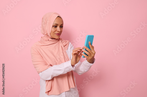 Isolated portrait of confident Arab Muslim middle aged woman in strict religious outfit and covered head in pink hijab texting a message on a mobile phone in her hands, colored background, copy space © Taras Grebinets