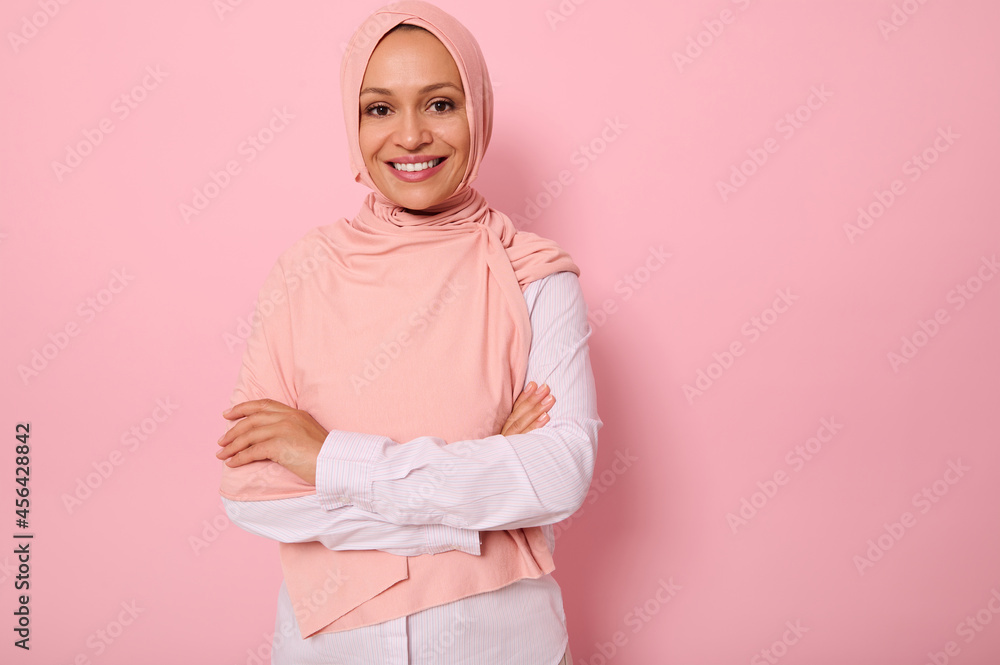 Fototapeta premium Waist-length portrait of a gorgeous Arabic Muslim woman in pink hijab posing looking to camera with attractive gaze, toothy smile, confident look and crossed arms, on pink background with copy space