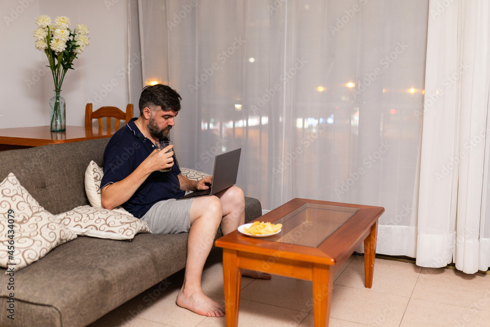 bearded man on sofa uses laptop and drink a beer in dining room, night