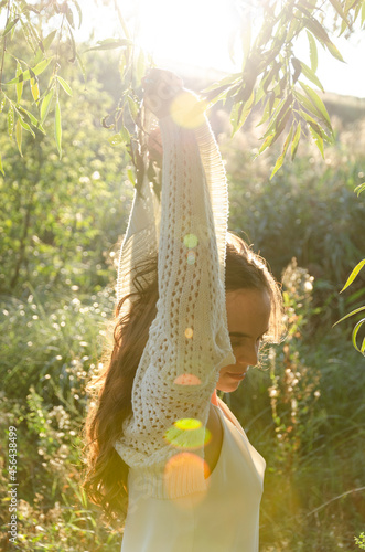 A young brunette woman is taking off her sweater outdoors, enjoying herself during golden hour. Backlit portrait.