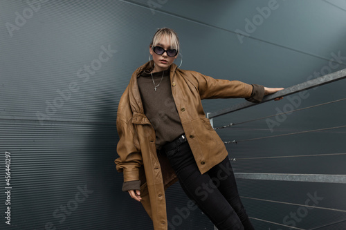 Stylish cool young woman model with fashionable sunglasses in an oversized leather jacket and a sweater with black jeans posing near a metal wall in the city