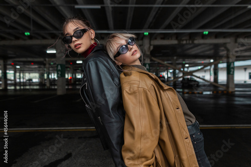 Two nice young model girls in youth style clothes with leather jacket with black jeans and vogue sunglasses posing in a parking lot in the city