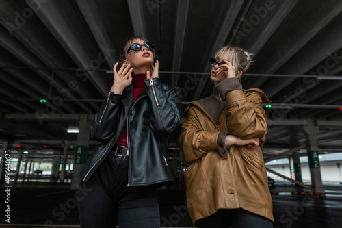 two beautiful young girls in youth style with leather jacket and black jeans with sunglasses posing in a parking lot on the street