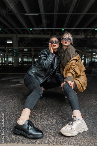 Stylish model girls in fashionable clothes with vintage leather jackets in black jeans with shoes posing on a parking in the city © alones