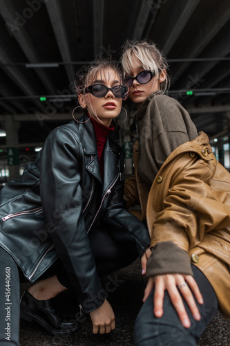 Trendy two fashion beautiful young women with stylish sunglasses in vintage leather jacket posing in the city