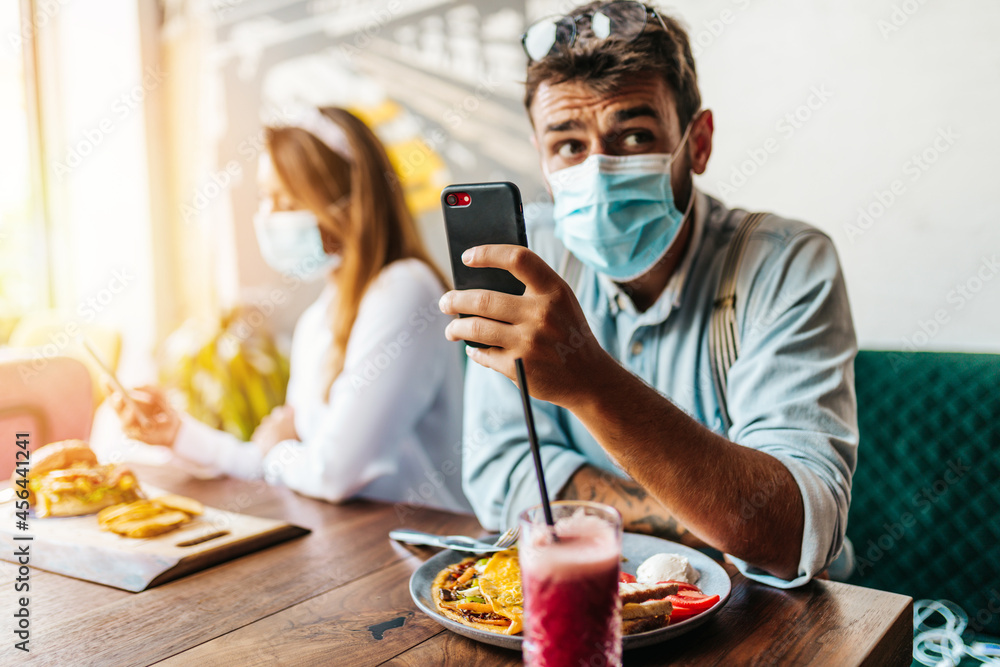 Young heterosexual couple with face protective masks sitting in modern restaurant. Coronavirus, food and drink concept. Young heterosexual couple with face protective masks sitting in modern restaurant. Coronavirus, food and drink concept.
