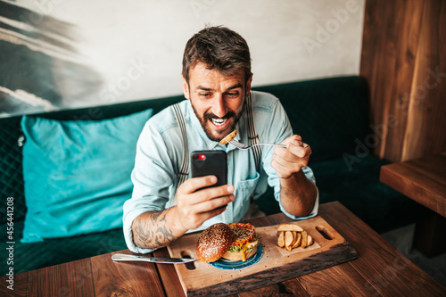 Handsome middle age man sitting in restaurant and enjoying in delicious burger. He is happy and smiled and uses his smart phone to talk with someone.