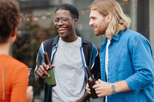 Waist up shot of two carefree young men drinking beer while enjoying outdoor party with friends