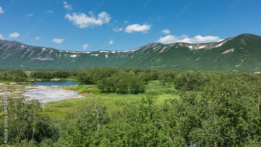 Fototapeta premium The caldera of an extinct volcano is surrounded by a mountain range. In the valley there is a blue lake, a green meadow, thickets of deciduous trees. Clouds in the azure sky. Summer. Kamchatka. Uzon