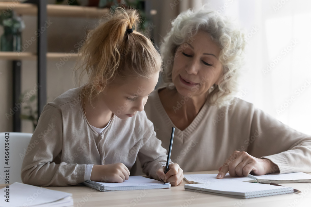 Caring middle aged old grandmother or nanny helping little girl grandchild with homework, teaching writing in copybook or explaining subject sitting together at table at home, homeschooling concept. Caring middle aged old grandmother or nanny helping little girl grandchild with homework, teaching writing in copybook or explaining subject sitting together at table at home, homeschooling concept.