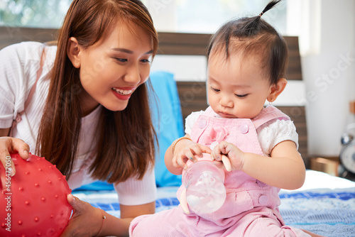 Smiling pretty Asian woman looking at daughter playing with feeding bottle on bed