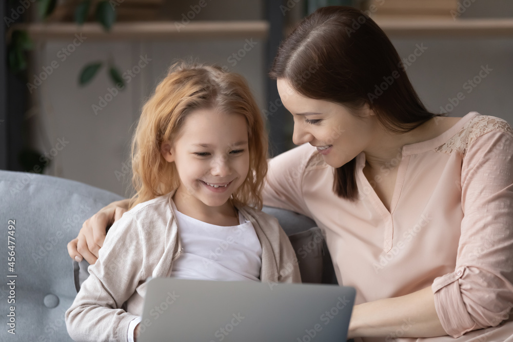 Caring young mother teaching little cute kid daughter using computer software apps, resting on comfortable sofa at home. Addicted to modern technology bonding two generations family playing online. Caring young mother teaching little cute kid daughter using computer software apps, resting on comfortable sofa at home. Addicted to modern technology bonding two generations family playing online.