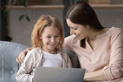 Caring young mother teaching little cute kid daughter using computer software apps, resting on comfortable sofa at home. Addicted to modern technology bonding two generations family playing online.