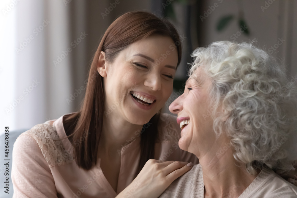 Head shot laughing attractive young woman cuddling smiling older retired mother, talking speaking communicating sharing life events at home, enjoying carefree leisure weekend pastime together. Head shot laughing attractive young woman cuddling smiling older retired mother, talking speaking communicating sharing life events at home, enjoying carefree leisure weekend pastime together.