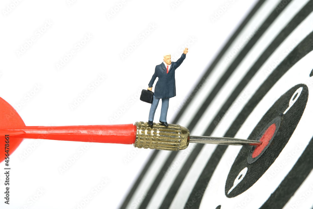Fototapeta premium Miniature people toy figure photography. A businessman standing above red arrow going to the center of dartboard while raise his hand. Isolated on white background.