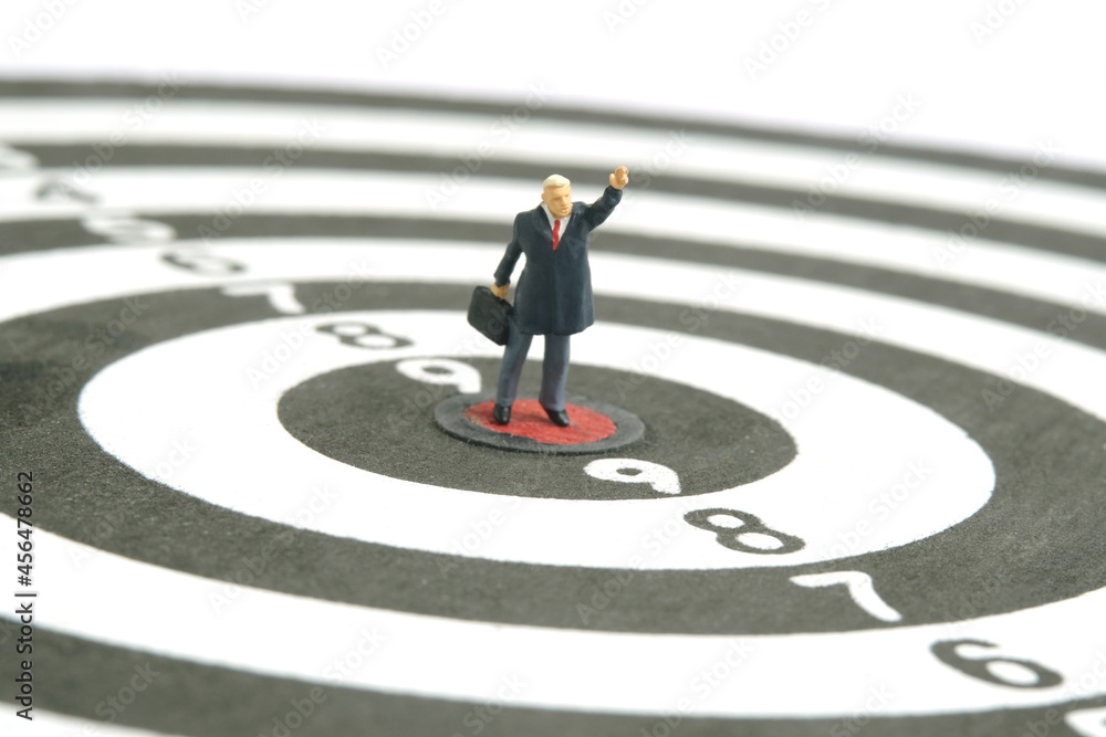 Miniature people toy figure photography. A businessman standing the center of dartboard while raise his hand. Isolated on white background. Miniature people toy figure photography. A businessman standing the center of dartboard while raise his hand. Isolated on white background.
