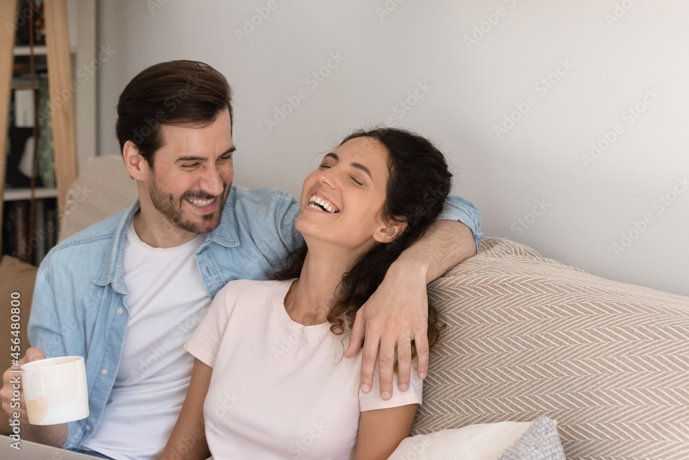Cheerful couple in love drink tea laughing hugging relax on sofa at home. Boyfriend girlfriend enjoy sweet tender moments together and morning coffee. Family weekend leisure at new own or rented house