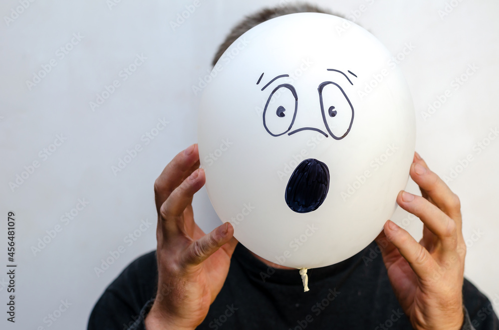 A man is holding a balloon with his face painted on it. The balloon is at head level. Unrecognizable personality. Emotion of surprise, indignation. Indoors. A man is holding a balloon with his face painted on it. The balloon is at head level. Unrecognizable personality. Emotion of surprise, indignation. Indoors.