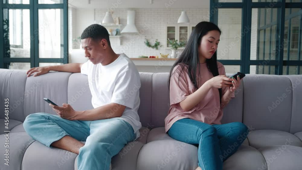 Young upset interracial couple of African American man and Asian woman sitting on the couch back to back, after the quarrel, holding smartphones in hands and turning away from each other. Young upset interracial couple of African American man and Asian woman sitting on the couch back to back, after the quarrel, holding smartphones in hands and turning away from each other.