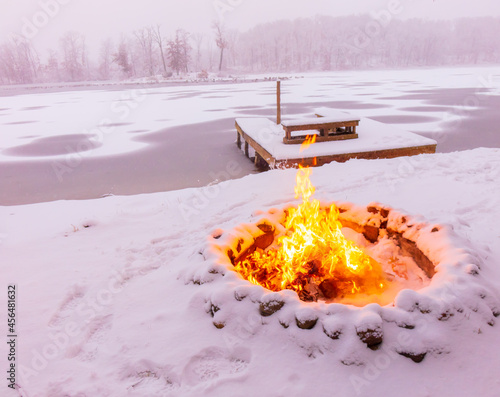 fire pit of stones with a roaring fire along a winter lake shoreline with background dock in ice and snow in January, Michigan, USA
