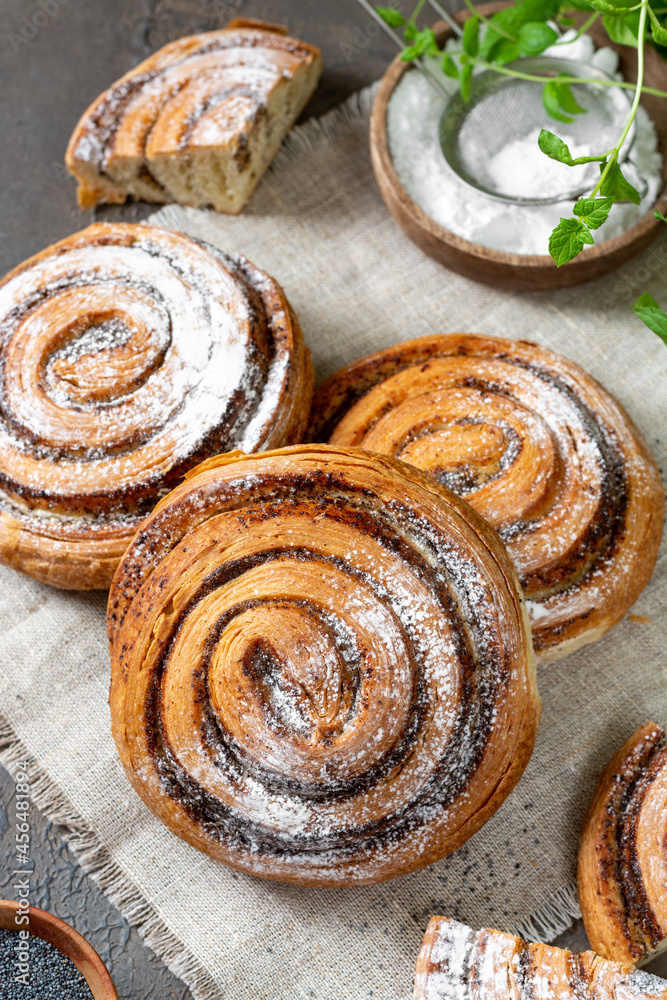 Sweet spiral bun with poppy seeds and powdered sugar large on a dark background. Delicious yeast pastries close-up. The concept of homemade high-calorie pastries Sweet spiral bun with poppy seeds and powdered sugar large on a dark background. Delicious yeast pastries close-up. The concept of homemade high-calorie pastries