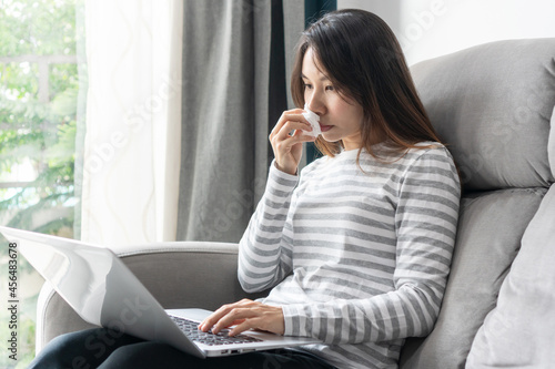 Allergic Asian woman blowing nose in tissue sit on sofa at home study or work on laptop, sick girl got flu caught cold having allergy symptoms holding napkin on hand while working on computer