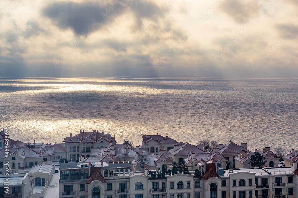 View of houses and rooftops against the backdrop of the sea in cloudy weather, the sun's rays make their way through the dramatic dark clouds over the sea