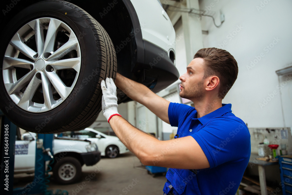 Cheerful, handsome, and confident car repair specialist in overalls repairs and replaces old parts with new ones in a car on a lift in service Cheerful, handsome, and confident car repair specialist in overalls repairs and replaces old parts with new ones in a car on a lift in service