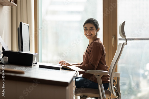Modern day worker. Portrait of happy biracial business woman freelancer sit by computer at comfy workplace at corporate workspace or at home. Smiling young indian lady office employee look at camera photo