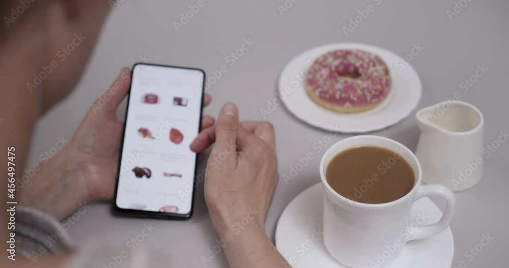 Woman at Home Sitting at Table With Coffee Donut Using Smartphone Buys in Internet Shop. Woman Orders Food Home In An Online Store Using Smartphone. Female Selects Vegetables in Grocery Online Store. Woman at Home Sitting at Table With Coffee Donut Using Smartphone Buys in Internet Shop. Woman Orders Food Home In An Online Store Using Smartphone. Female Selects Vegetables in Grocery Online Store.