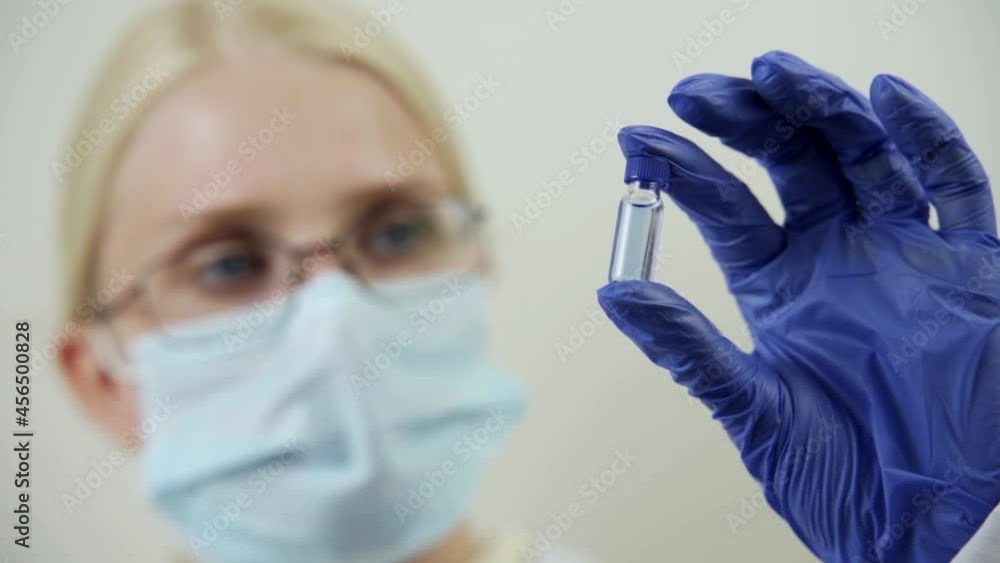 A serious doctor in a medical mask and rubber gloves holds an ampoule with a blue drug in his hands. Science and medicine. Close-up, slow motion, blurred background, HD. A serious doctor in a medical mask and rubber gloves holds an ampoule with a blue drug in his hands. Science and medicine. Close-up, slow motion, blurred background, HD.