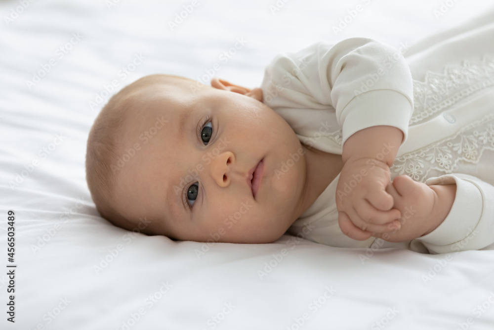 Little adorable few month baby lying on mattress, looking at camera. Cute infant boy or girl in bodysuit, resting in bed with white linen. Family, childhood, childbirth concept. Child portrait Little adorable few month baby lying on mattress, looking at camera. Cute infant boy or girl in bodysuit, resting in bed with white linen. Family, childhood, childbirth concept. Child portrait