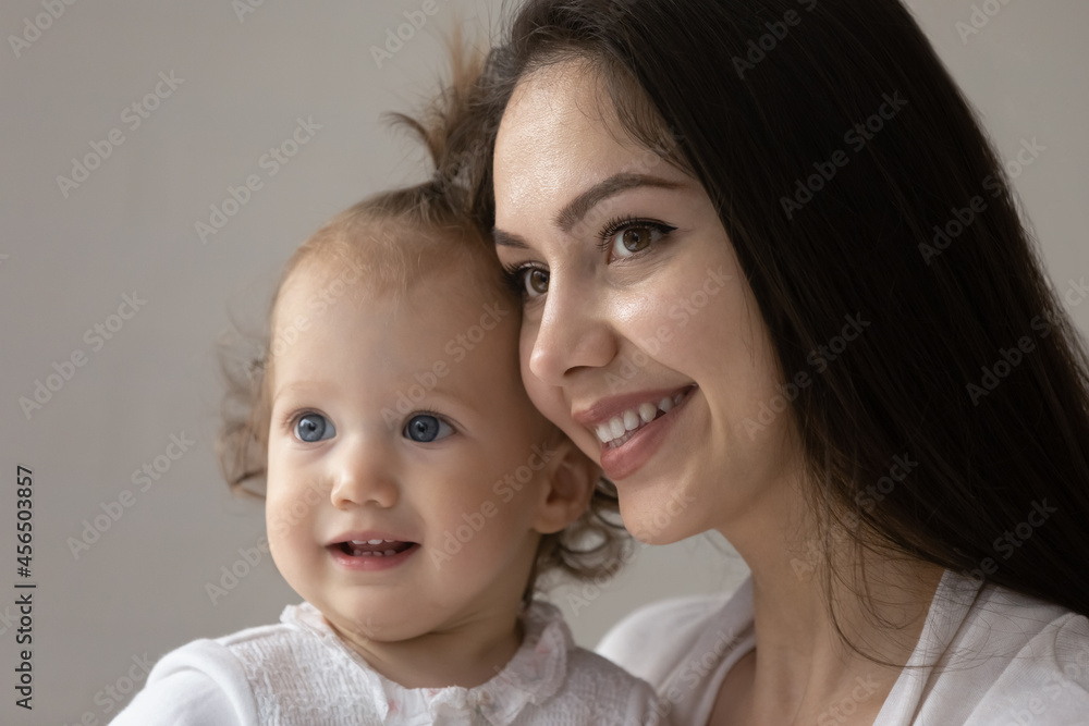 Close up portrait of happy new mom and cute blue eyed baby girl enjoying time together. Smiling young mother holding adorable toddler kid in arms, looking away. Motherhood, child care concept Close up portrait of happy new mom and cute blue eyed baby girl enjoying time together. Smiling young mother holding adorable toddler kid in arms, looking away. Motherhood, child care concept