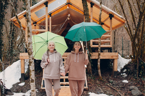 Elderly and young adult women with umbrella at glamping camping tent. Modern autumn vacation lifestyle concept