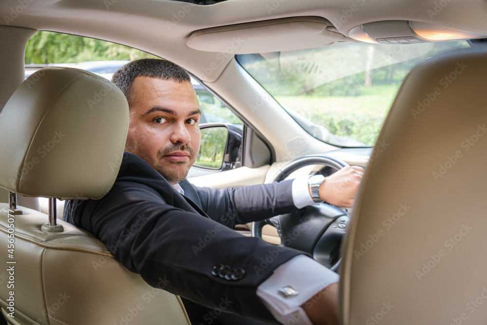 Serious young man of Middle Eastern appearance in a business suit at the wheel an expensive car. A man inspects a car before buying. Car sales manager. Man turned around from the front seat of the car