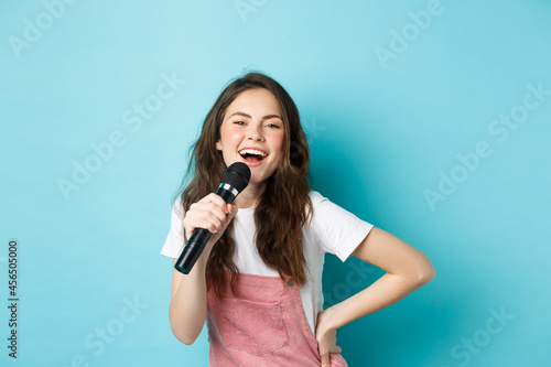 Fotografie, Obraz Cheerful young woman singing karaoke, holding microphone and smiling, having fun