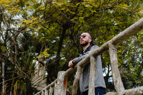 young hispanic latino man smiling leaning on a railing in a park outside, in autumn, wearing sportswear and sunglasses. copy space