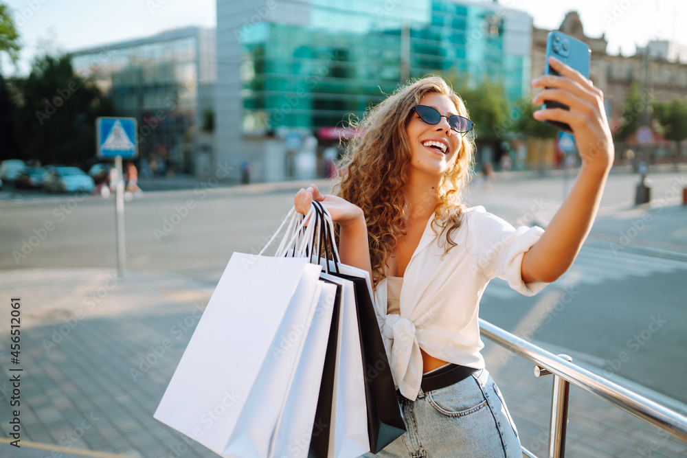 Beautiful woman  with shopping bags makes selfie. Young woman walking around the city after shopping. Black friday, sale, consumerist, lifestyle concept.