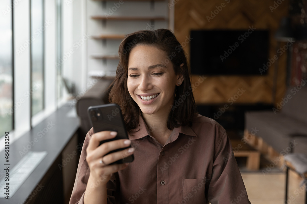 Fototapeta premium Smiling millennial Caucasian woman look at smartphone screen talk speak on video call online. Happy young female use cellphone have webcam digital virtual event on gadget. Communication concept.