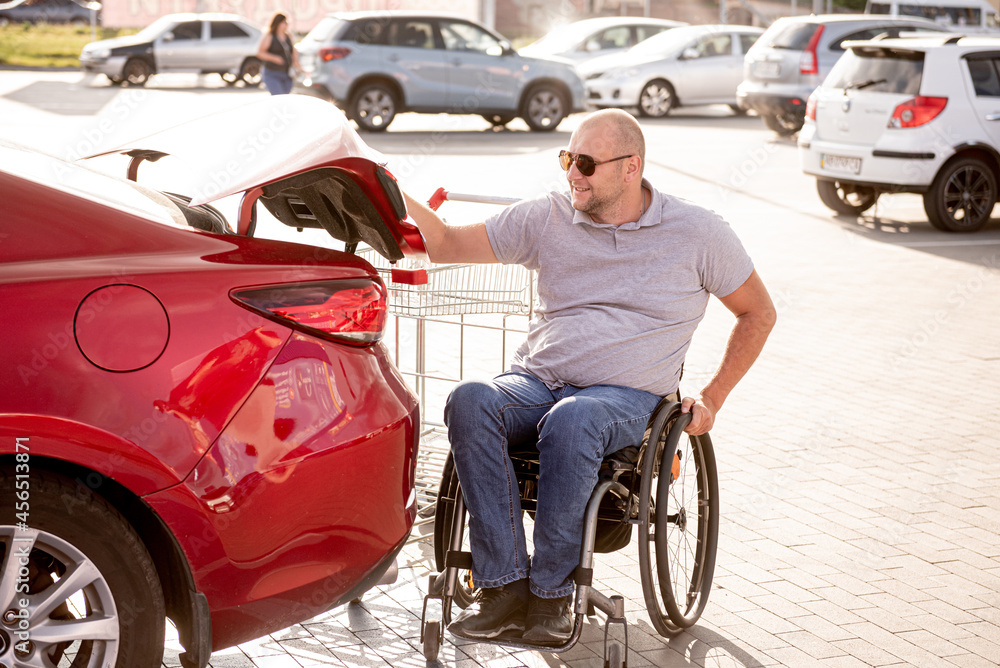 Adult disabled man in a wheelchair puts purchases in the trunk of a car in a supermarket parking lot Adult disabled man in a wheelchair puts purchases in the trunk of a car in a supermarket parking lot