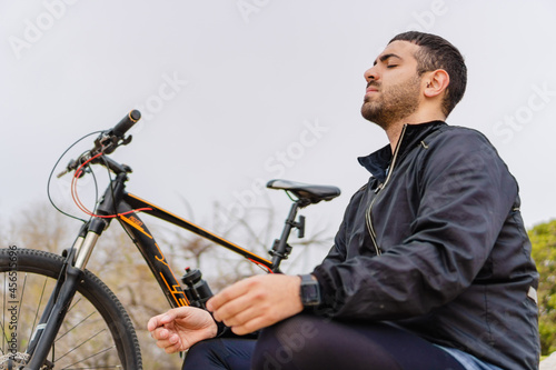 Latino man sitting in the park with his bicycle next to him meditating with his eyes closed.
