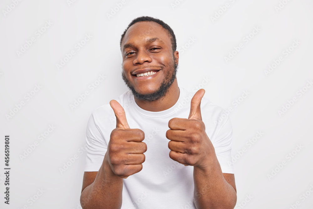 Happy smiling dark skinned man approves incredible promo keeps thumbs up likes and agrees being satisfied with something wears casual clothes praises cool job isolated over white background. Happy smiling dark skinned man approves incredible promo keeps thumbs up likes and agrees being satisfied with something wears casual clothes praises cool job isolated over white background.