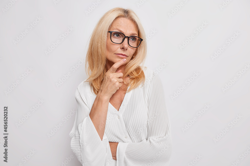 Indoor shot of thoughtful light haired woman of middle age recalls something from life looks away keeps hand on chin makes up plan wears spectacles for vision correction stylish silk white blouse Indoor shot of thoughtful light haired woman of middle age recalls something from life looks away keeps hand on chin makes up plan wears spectacles for vision correction stylish silk white blouse