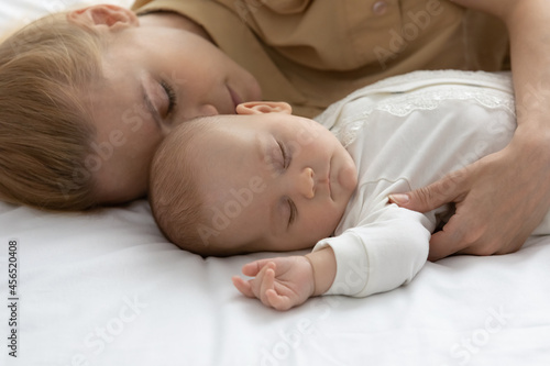 Peaceful tired mother and calm infant sleeping together, lying in bed on white linen, sheet, relaxing in bedroom, taking break. Pretty new mom relaxing near sleepy baby at home. Child care, motherhood
